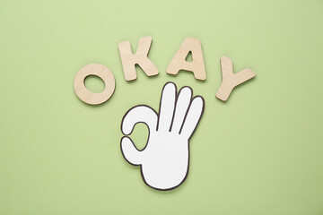 Fototapeta na wymiar Word Okay made of wooden letters and paper cutout (OK hand gesture) on pale olive background, flat lay