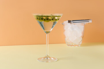 Glass of martini with olives, ice and tongs on color background