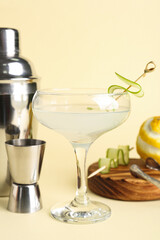 Glass of tasty martini, shaker, jigger and lemon on light yellow background