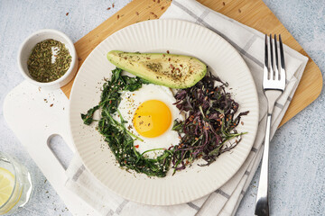 Plate with tasty fried egg, avocado and greens on light background