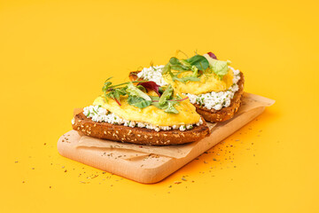 Wooden board with tasty scrambled eggs sandwiches and greens on yellow background