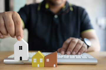 Man use the keyboard for searching a home and choose a mini wood house model from the model on a wood table. Planning to buy property. Concept of choosing the best home. Family fulfillment.