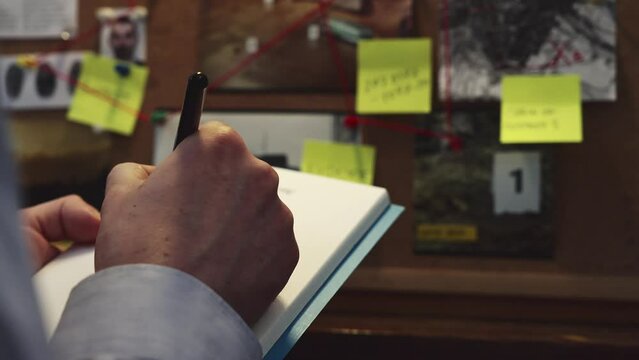 In front of the evidence board, the detective captures important details by taking notes in a notebook