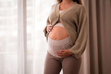 Pregnant woman wearing comfortable, orthopedic belly belt, bandage, touching baby bump near window, cropped shot,Pastel colours.Healthy pregnancy,backaache,pain, support concept.Copyspace.