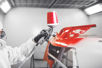 Spray painter worker in protective glove with airbrush pulverizer painting red car bumper in white paint chamber.