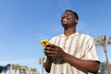 Happy black man laughing outdoors using mobile phone. Blue sky background. Copy space.