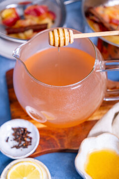 Honey being dripped into apple tea in a glass tea pot, cloves, apple slices around it.