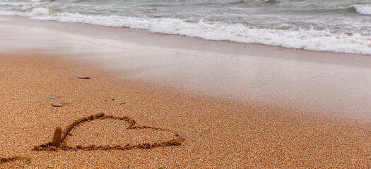 The seas are full on the sandy shore on a cloudy day. A heart is painted on the sand, and the waves wash away the drawing. Rest on the sea. There is an empty space for text