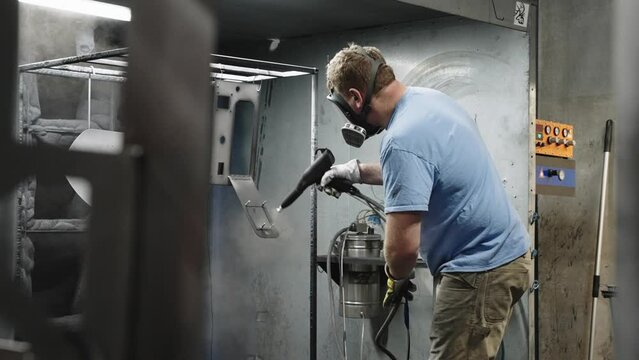 Blue collar worker spray painting metal with powder coating in an industrial room, slow motion