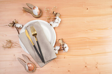 A top view on a Empty white ceramic plate, golden cutlery on linen napkin, bunny-shaped egg holders, gold foiled eggs, cotton flower decoration on a wooden background, copy space, top view