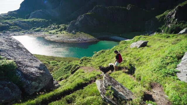 Female Trekker Walking Downhill With Pet Dog With Stunning Scenery Of Turquoise Blue Mountain Lake In Lovund, Norway. high angle, pan left