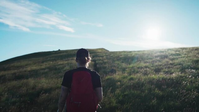 Following A Woman Wearing Cap And Backpack While Trekking On Grassy Hill With Pet Dog On A Sunny Day. medium shot