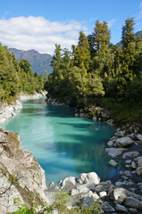 Fototapeta premium Glacier-fed river gorge lined with by quartz rocks and native rainforest bush under a blue sky in Hokitika, West Coast, New Zealand