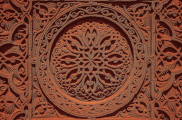 Fototapeta na wymiar Carved round ornament with intertwining strips forming a pattern placed in a round frame on a red volcanic stone. Traditional tufa stone carving ornament on a wall of Armenian Orthodox Church.