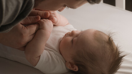 Happy father plays with cute baby in child crib in the morning, touches and kisses little hands. A...
