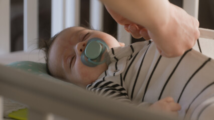 Face close up shot of sweet and beautiful newborn baby in white cradle. Father takes off bodysuit from sleeping infant with pacifier in mouth. Concept of parenthood, carefree childhood and family.