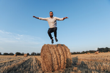Naklejka na ściany i meble Wedding photo. Stylish groom in white shirt jumping down from hay bale and looking at camera. Bearded man. Style. Emotions
