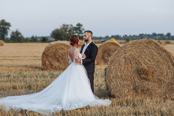 Naklejka na ściany i meble The bride and groom are standing in the field, and behind them are large sheaves of hay. The bride stands with her shoulders turned to the camera. Long elegant dress. Stylish groom. Summer