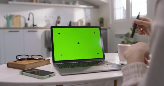 Laptop with green screen on table against background of kitchen set. Template to insert. Woman is sitting at desk, working on laptop. Female hands and fingers are gesticulating and twisting black pen