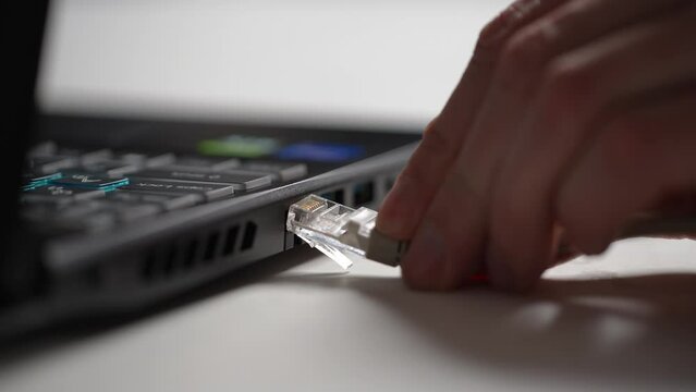 Close-up cropped shot of unrecognizable man inserting internet cable RJ 45 connections to laptop computer at table. Detail male hands of ethernet cable plug inserted into port on side of laptop.