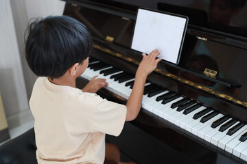 Kid asian black hair boy sitting and playing piano with tablet in living room house indoor. Musical and relaxation makes them happiness. Health care lifestyle concept.