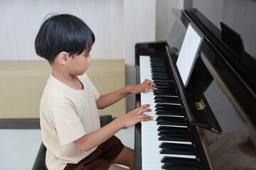 Kid asian black hair boy sitting and playing piano with tablet in living room house indoor. Musical and relaxation makes them happiness. Health care lifestyle concept.