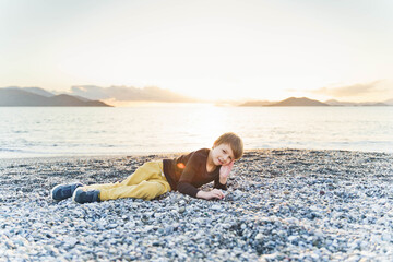 Little boy lies on a pebble beach at sunset.