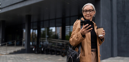 a mature adult female director with gray hair during a break with a phone stands against the backdrop of a business center
