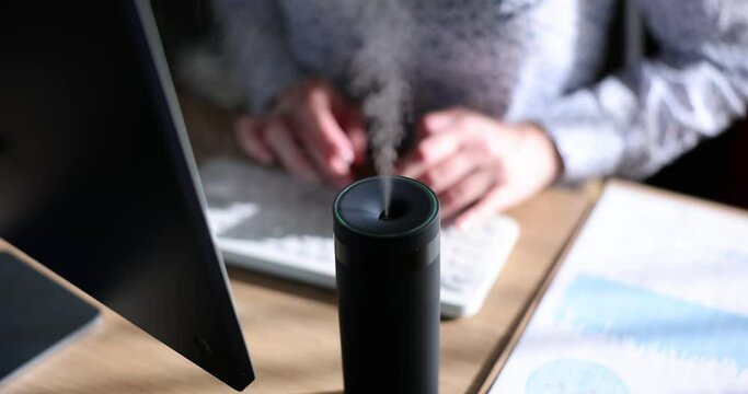 Freelancer types article on keyboard inhaling scent of essential oils. Modern diffuser spreads aroma and humidifies air in room slow motion