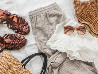Women's clothing, comfortable vacation clothes - linen trousers, sandals, straw basket, blouse and sunglasses on a light background, top view