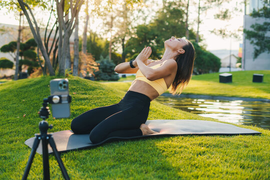 Adult woman blogger in sportswear recording video on camera phone as she doing yoga on mat in summer park outdoors. Training workout. Healthy lifestyle, sport