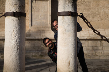 Young Spanish gay male couple have fun among the marble columns and chains in the city. They are on their honeymoon vacation. The couple are married. Concept of gay rights