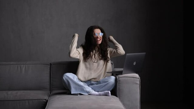 Young woman in glasses working on laptop get winning news and gesture while sit on sofa at home