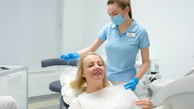 nurse in dental clinic puts on woman bib napkin for patient in dental white background of latest technology excellent light doctor nurse patient preparing for procedure pull up glass table with tools