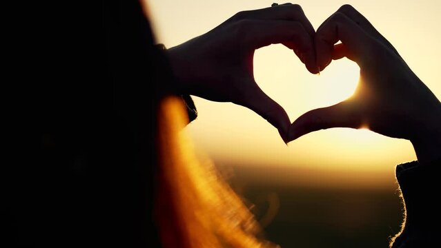 Happy girl in park at sunset. Finger shaped heart shape. Hands of girl shape of heart. Summer dream. Happiness of freedom in a field at sunset.Sunlight between fingers.Silhouette of happy girl in park