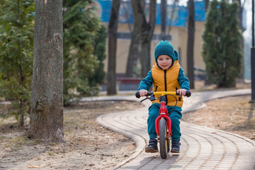 Fototapeta premium A cheerful little boy rides a bicycle outdoors. A happy child walks in the spring park. The baby is dressed in a fashionable yellow vest and turquoise jumpsuit.