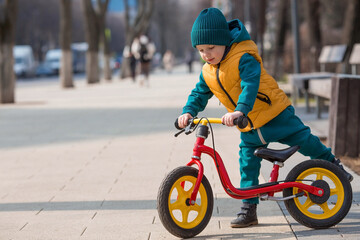 A cheerful little boy rides a bicycle outdoors. A happy child walks in the spring park. The baby is dressed in a fashionable yellow vest and turquoise jumpsuit. - obrazy, fototapety, plakaty