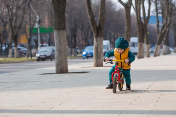 A cheerful little boy rides a bicycle outdoors. A happy child walks in the spring park. The baby is dressed in a fashionable yellow vest and turquoise jumpsuit.