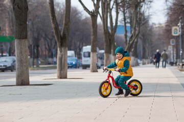 A cheerful little boy rides a bicycle outdoors. A happy child walks in the spring park. The baby is dressed in a fashionable yellow vest and turquoise jumpsuit.