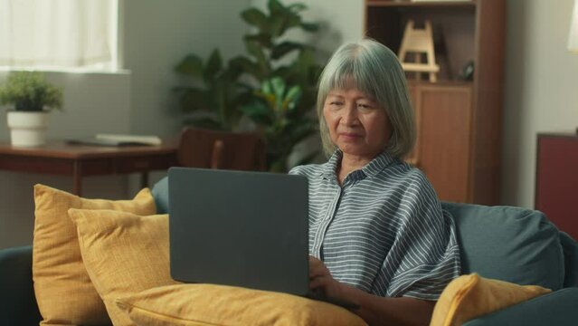Beautiful elderly Asian woman using laptop texting chatting working on notebook computer. Old female using technology in daily life sit on couch in cozy living room. Senior Lifestyle Concept.