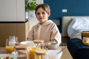 Obraz na płótnie Canvas Portrait of happy small european boy having breakfast at home. Smiling little child schoolboy sitting at kitchen table in morning. Interested kid boy looking at camera eating cornflakes before school