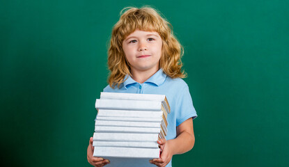 Child holding stack of books with mortar board on blackboard. Back to school. Schoolchild in class. Happy kid against green blackboard. Education and creativity concept