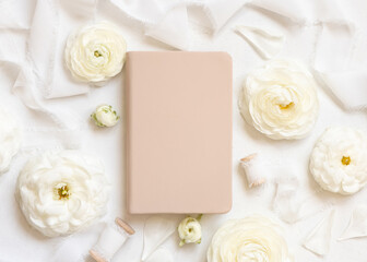 Obraz na płótnie Canvas Cream hardcover textbook near cream roses and white silk ribbons top view, mockup