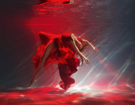 fashion model posing with sexy legs. Fantasy woman dancing under water sea, red long silk dress fabric floating. Belerina fairy girl dancer swims in dark deep pool underwater shooting Art Magic light 