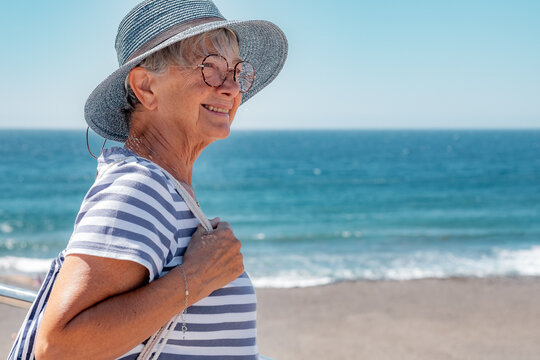 Smiling caucasian senior woman in outdoors summer sea vacation. Smiling elderly lady with hat dressed in blue expressing good mood and joy. Horizon over water