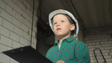 A little boy construction worker in a white helmet and a green shirt with a tablet with documents in his hands stands on the construction site and looks into the distance. High quality photo