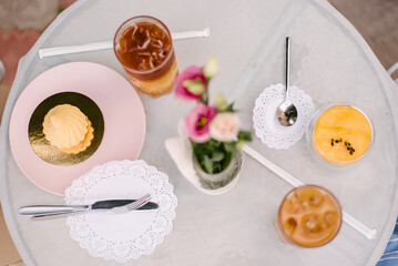 There are desserts on a table with a white tablecloth. Nearby are glasses of drink with ice and kitchen utensils. Table on the summer terrace of the cafe