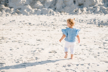 Kid runs. Child baby barefoot walk on the sand on the sandy beach. Little girl toddler playing and walking in the sand. The concept of family summer vacation with children outdoors. Back view.