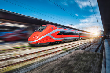 Obraz na płótnie Canvas High speed train in motion on the railway station at sunset. Fast red modern intercity train and blurred background. Railway platform. Railroad in Italy. Commercial. Passenger railway transportation