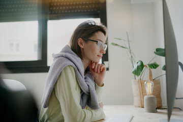 A young woman working on a desktop computer in a bright and sunny space fully immersed in her work. The sunlight through the window, casting a warm glow on her workspace. 